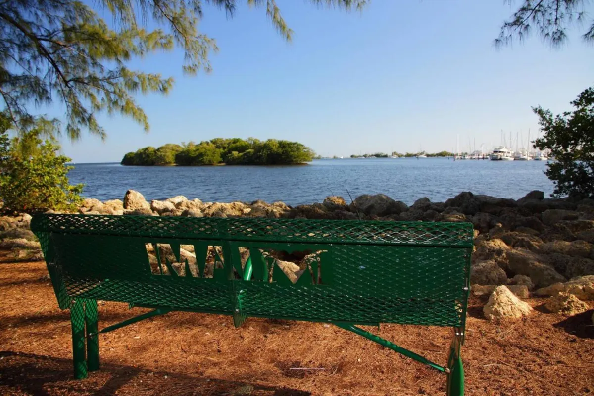 A peaceful waterfront view featuring a green bench with a 'MIAMI' cutout, calm water, and an island in the distance. The scene invites thoughts of picnics, quiet moments, and easy access to local parks and outdoor attractions.