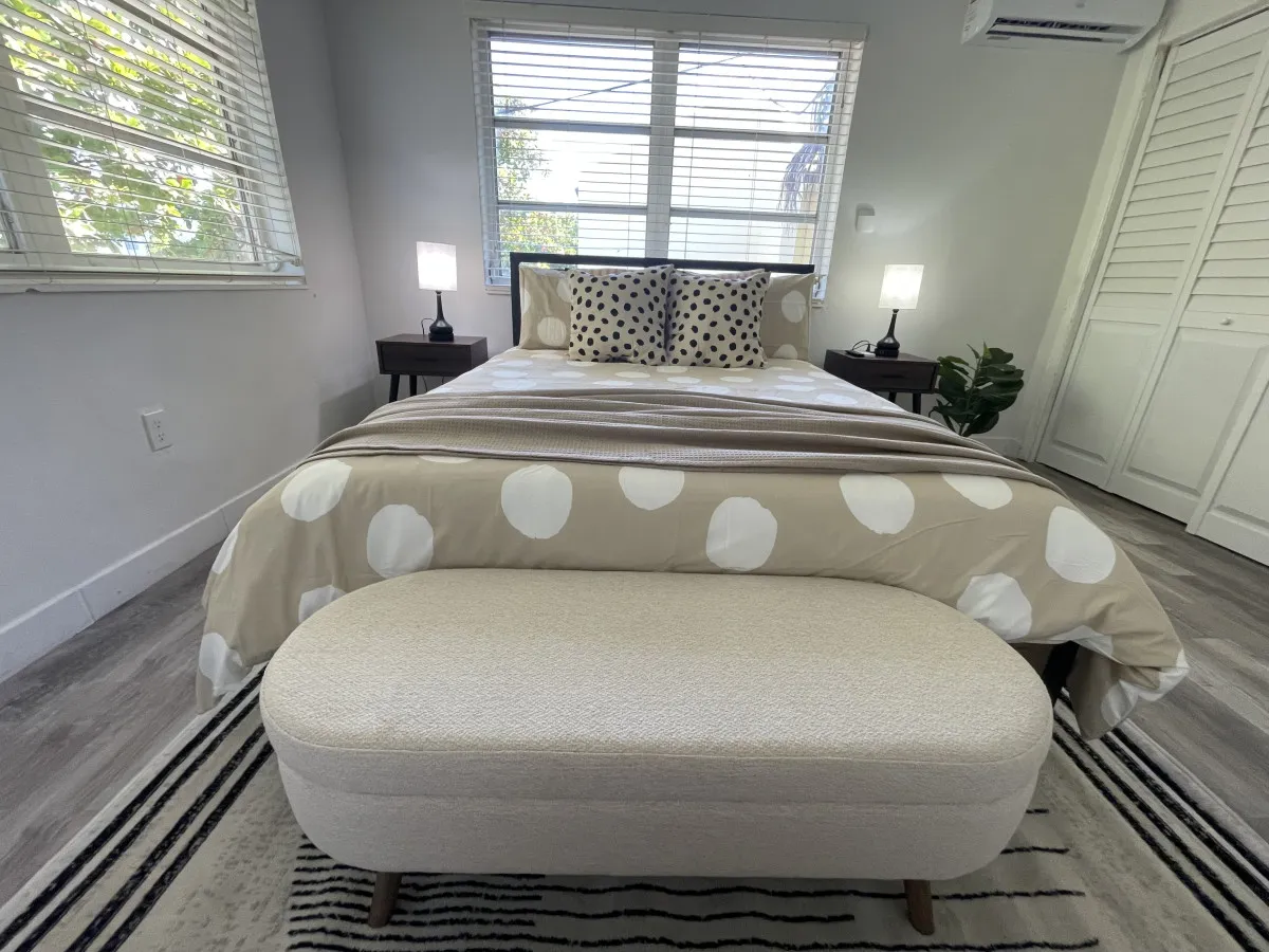 This image showcases a serene bedroom featuring a queen size bed adorned with stylish polka dot bedding, bathed in natural daylight from a large window. Matching bedside tables with lamps and a plush upholstered bench at the foot of the bed add to the inviting, cozy atmosphere.