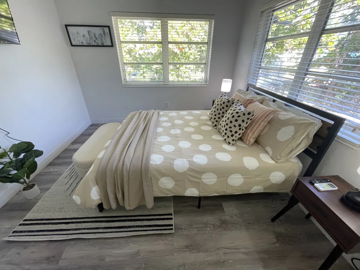 A calm and comfortable bedroom view with a queen-size bed dressed in polka dot bedding, expansive corner windows with white blinds, a small wooden bedside table with a lamp and remote, and a potted plant for a modern touch. The smart TV is visible, reinforcing the room’s functional amenities.