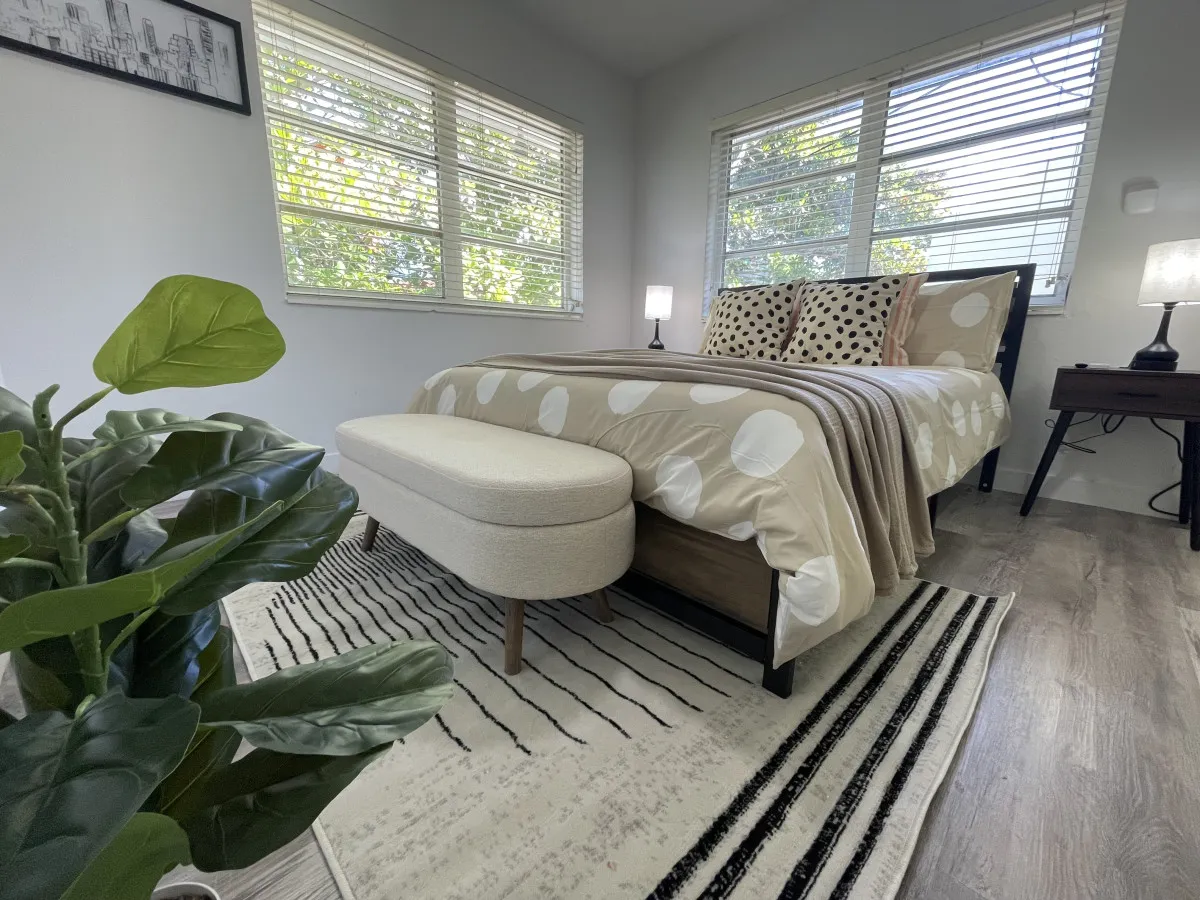 This image shows a serene bedroom featuring a queen-size bed with polka dot bedding, large windows with white blinds that let in abundant natural light, a modern bench at the foot of the bed, and a green leafy plant in the foreground adding a touch of freshness. The space is airy and inviting, ideal for relaxation and restful nights.