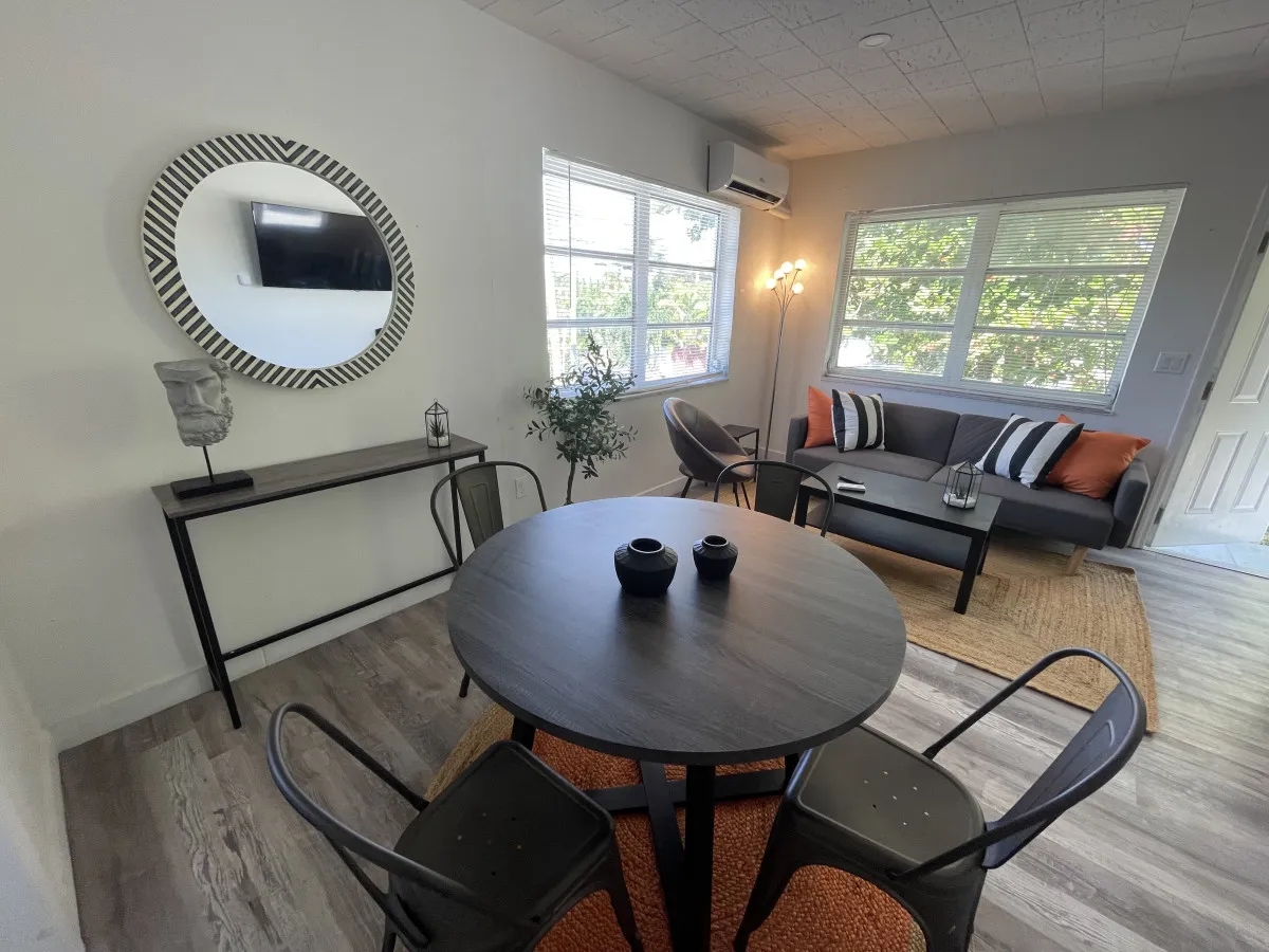 A sunlit, modern living/dining space featuring a round black dining table with metal chairs, a gray sofa adorned with striped cushions, a patterned decorative mirror, and a contemporary floor lamp. The open-plan layout and artistic touches create a welcoming, stylish ambiance perfect for relaxing after a day in the neighborhood.