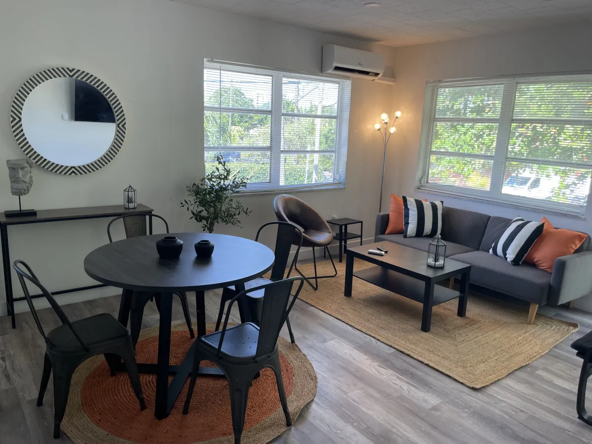 A view of the modern living and dining area featuring a gray sofa with colorful pillows, a round black dining table set with four metal chairs, a decorative mirror, and a multi-bulb floor lamp. The space feels airy and inviting, with subtle hints of natural light from the window, suggesting a connection to the outdoors and the urban environment.