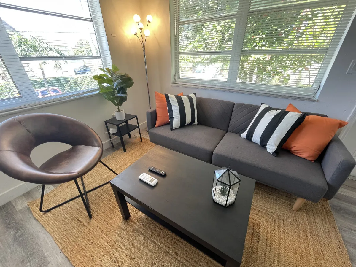 A cozy scene with a gray sofa adorned with colorful pillows, a round leather chair, a black coffee table with decor and remotes, and a corner standing lamp. The setup invites relaxation, watching TV, or enjoying coffee after a day in the city.