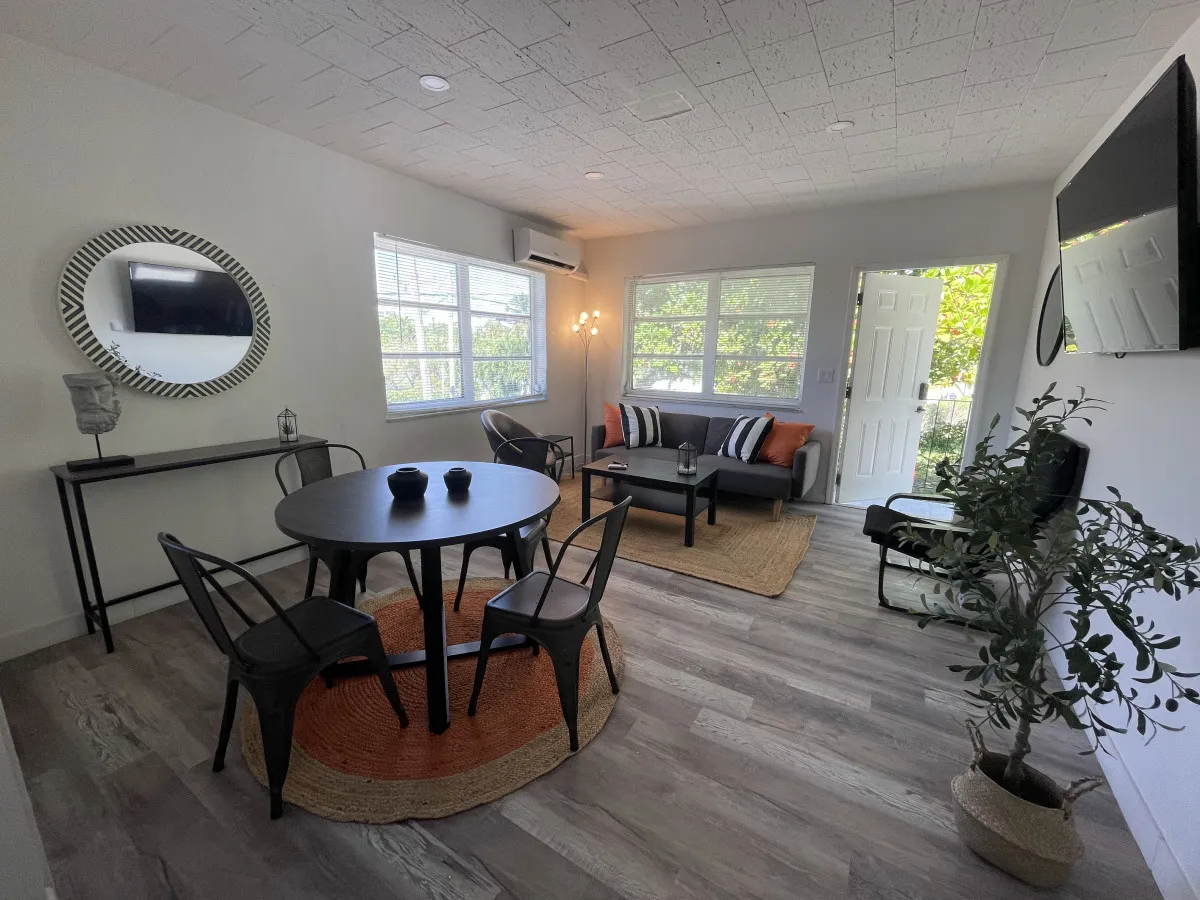 This image showcases a bright living room with a gray sofa adorned with orange and black striped cushions, a round black dining table with four metal chairs, a large wall-mounted smart TV, and a decorative round mirror with a black and white zigzag pattern. The space is filled with natural light from large windows, emphasizing the room's modern comforts and inviting atmosphere.
