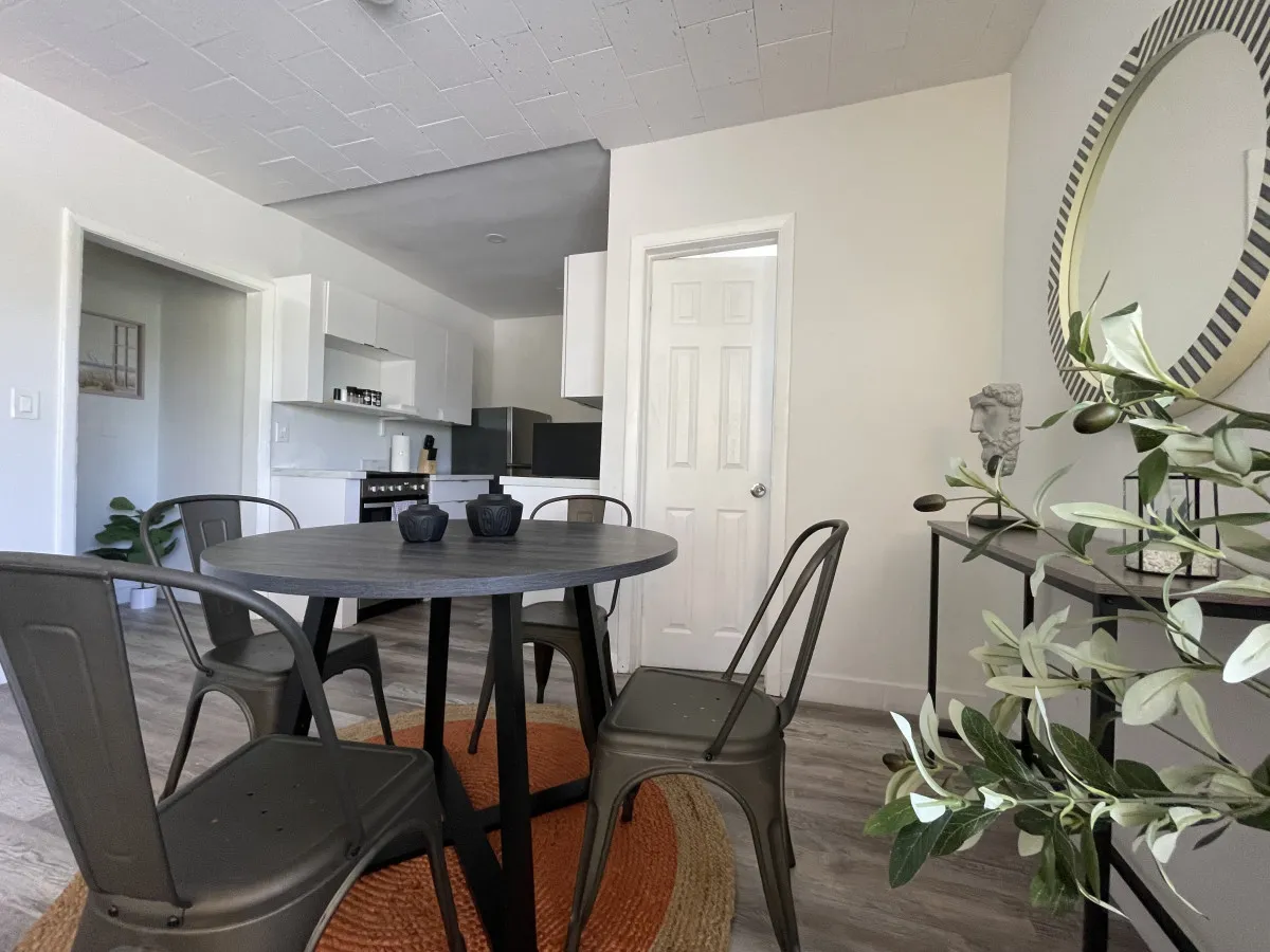 A cozy dining space featuring a round dining table with four metal chairs, set adjacent to a modern kitchen. The space is styled with a decorative round mirror, a green plant, and white cabinetry, creating an inviting spot for meals, socializing, or morning coffee.