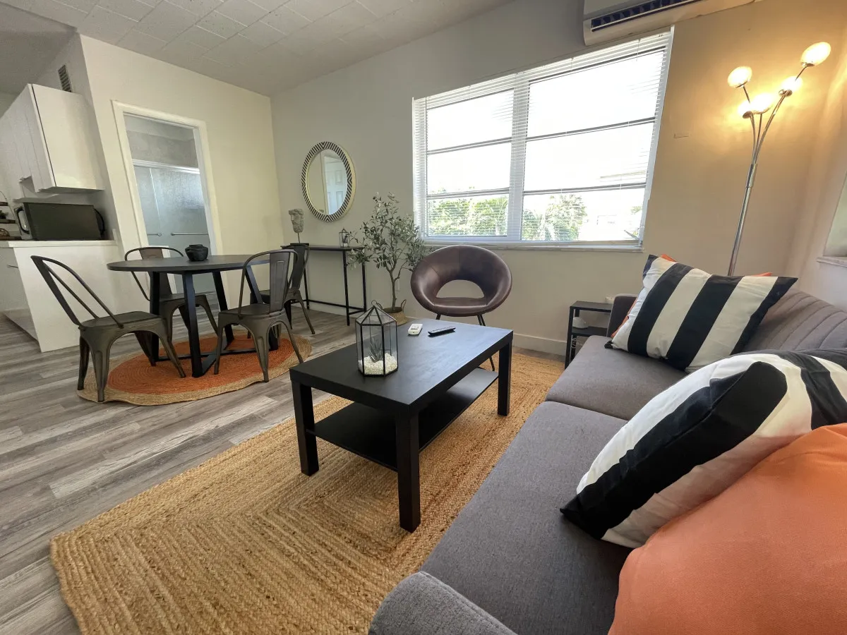A modern, comfortable living area with a gray sofa adorned with striped and solid pillows, a black coffee table, and a round dining table with metal chairs. Large windows let in abundant natural light, creating a casual, homely atmosphere ideal for relaxation or working.