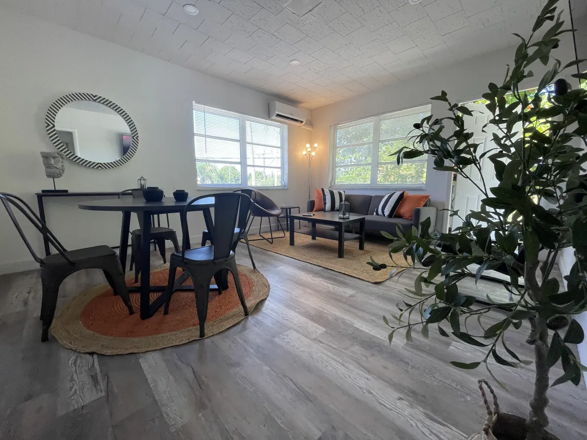 A welcoming living area featuring a modern gray sofa with striped and orange pillows, a round black dining table with metal chairs, large windows streaming in sunlight, a decorative mirror, and a floor lamp. The room feels open and stylish, perfect for unwinding or planning city adventures.