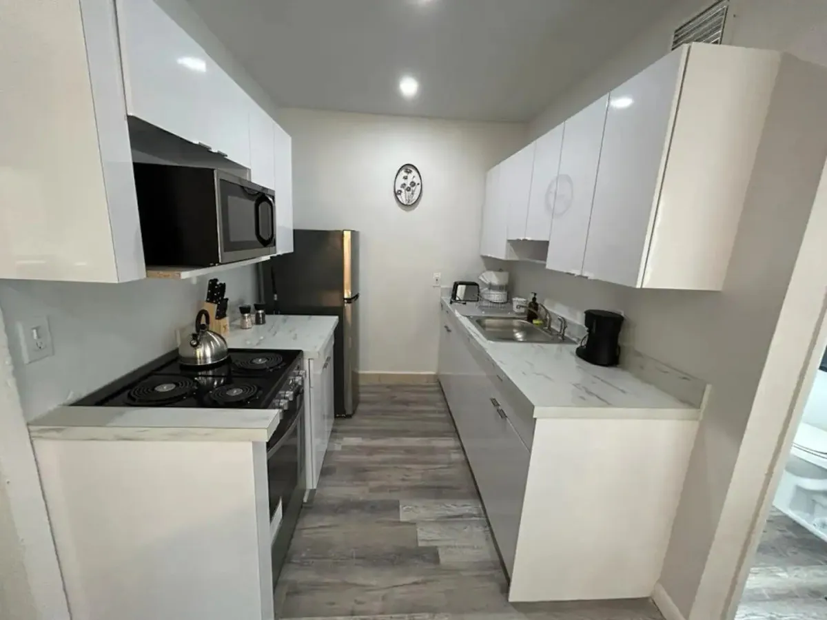 A bright, modern kitchen featuring white glossy cabinets, marble-patterned countertops, stainless steel appliances, and a variety of kitchen utensils and small appliances displayed on the counter. The space is organized and inviting, clearly ready for meal preparation or making coffee.