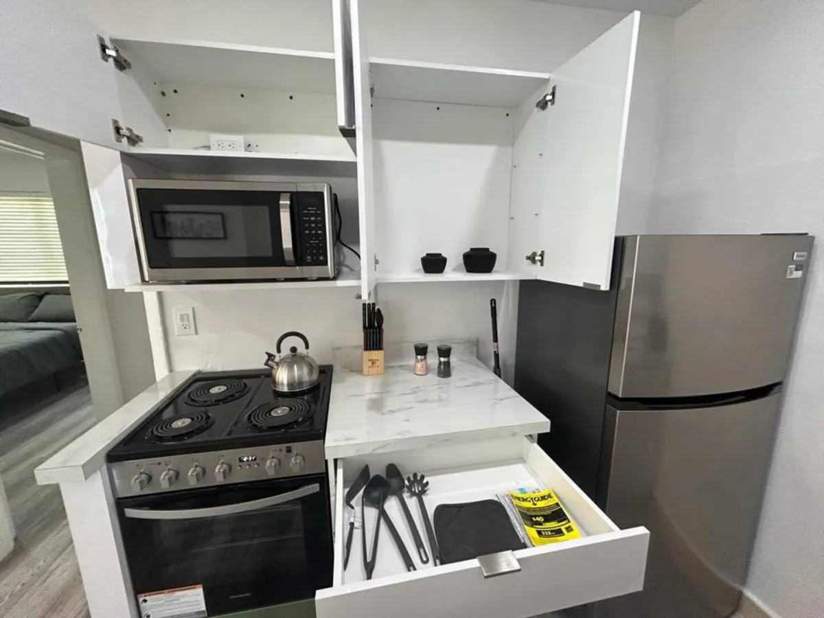A compact, efficient kitchen corner with an electric stove and kettle, open upper cabinets revealing minimal items, a drawer with cooking utensils, and a stainless steel refrigerator. The space is practical and organized, emphasizing the kitchen's functionality for cooking and storing groceries.