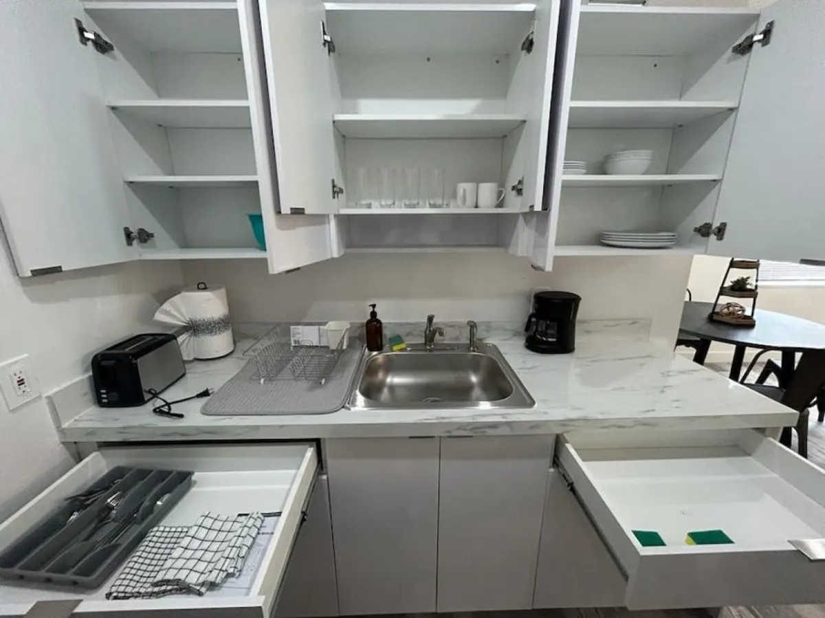 A practical kitchen space featuring a stainless steel sink, open cabinets stocked with dishes and glasses, a toaster and coffee maker, and a dish drying rack on the countertop. The setup is inviting and functional for daily use.