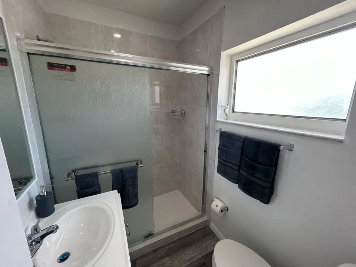 A modern bathroom featuring a frosted glass sliding shower door, white ceramic sink with chrome faucet, two dark towels on towel bars, and a frosted window bringing in natural light. The setup is neat, clean, and functional, with all essentials in view.