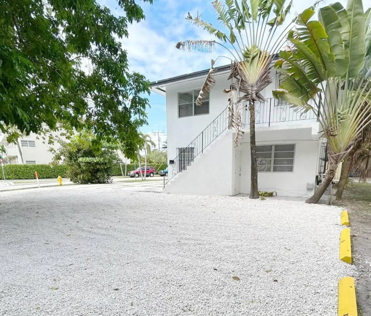 A white multi-family building with easy-access exterior stairs, a gravel parking lot with yellow stops, and lush tropical palm trees. The scene communicates convenience for vehicle parking and a welcoming atmosphere with greenery and shade, reflecting the urban Miami setting.