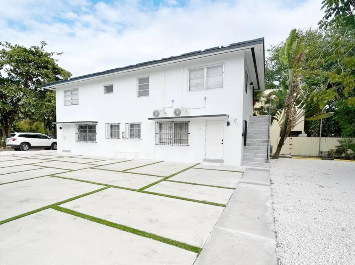 This image features the exterior of a white multi-family building with clearly visible exterior stairs and a convenient parking area. The surrounding greenery and trees add a touch of nature, while the functional setup highlights secure, on-premises parking and easy access points for residents and visitors alike.