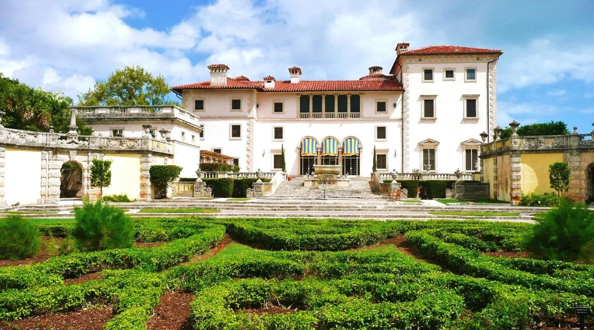 This image shows a grand Mediterranean-style mansion surrounded by a lush, well-manicured garden. The symmetrical greenery, central stone fountain, and arched windows with striped awnings evoke a sense of classic Miami elegance and outdoor leisure. The bright, inviting setting hints at the upscale and vibrant character of the Coconut Grove neighborhood.