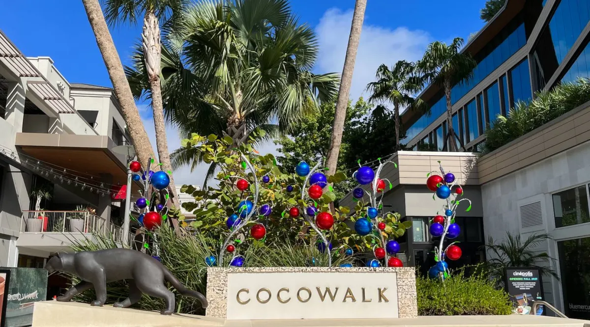 The energetic exterior of the CocoWalk area, featuring a prominent stone sign, colorful public art, and lush palm trees. Pedestrians can be seen enjoying the vibrant outdoor space, hinting at the lively city atmosphere and proximity to shopping, dining, and entertainment.